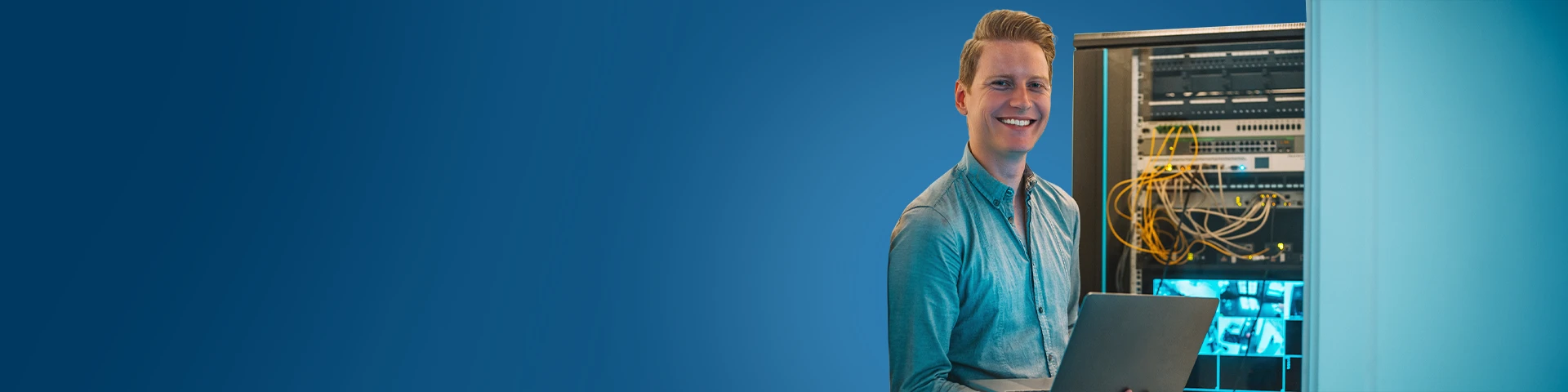 Un giovane uomo sorridente in camicia azzurra tiene in mano un computer portatile davanti a un rack di server aperto, con numerosi cavi di rete visibili.