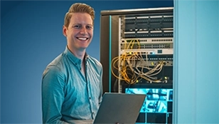 Un giovane uomo sorridente in camicia azzurra tiene in mano un computer portatile davanti a un rack di server aperto, con numerosi cavi di rete visibili.
