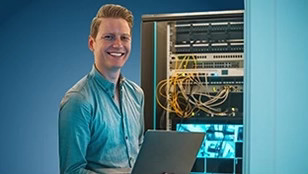 Un giovane uomo sorridente in camicia azzurra tiene in mano un computer portatile davanti a un rack di server aperto, con numerosi cavi di rete visibili.
