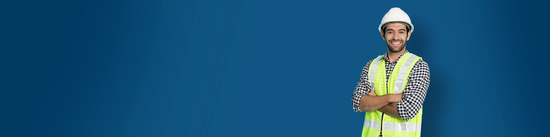 Un lavoratore edile sorridente indossa un casco bianco, un giubbotto di sicurezza giallo catarifrangente e una camicia a quadri. È in piedi con le braccia incrociate su uno sfondo blu scuro.