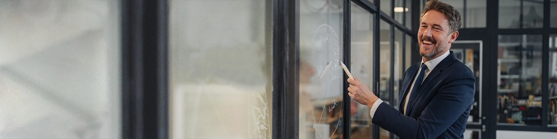 Uomo d'affari sorridente con la barba, che indossa un completo scuro e una cravatta, in piedi in un ufficio moderno con partizioni in vetro. L'uomo è girato verso destra, guardando verso una lavagna trasparente non visibile, e tiene un pennarello bianco nella mano destra come se stesse scrivendo o puntando qualcosa. La sua espressione è gioiosa e coinvolta. L'ambiente dell'ufficio sullo sfondo è leggermente sfocato ma mostra strutture in vetro e metallo.