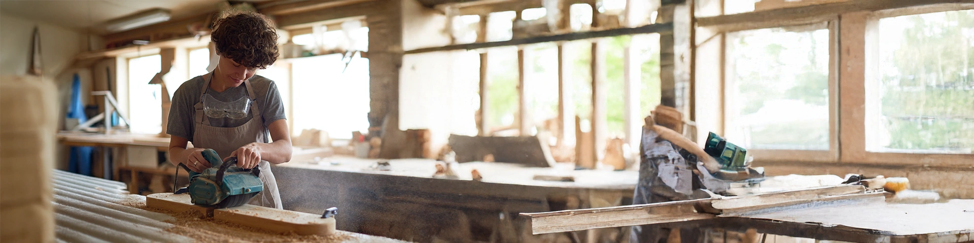 Primo piano di un giovane artigiano al lavoro all'interno di una falegnameria luminosa. Il ragazzo, che indossa occhiali protettivi trasparenti al collo e un grembiule grigio, è concentrato mentre utilizza una levigatrice elettrica su una lunga asse di legno appoggiata su un banco da lavoro. La luce naturale proviene dalle finestre sullo sfondo, piene di attrezzi e segatura.