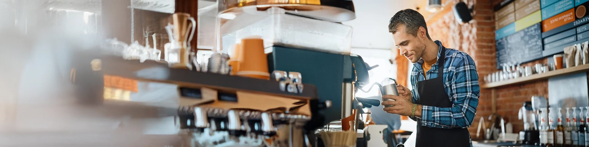 Interno di un bar moderno con pareti in mattoni a vista dove appare un barista. Lui è un uomo di mezza età, con capelli corti grigi e una camicia a quadri blu e bianca sotto un grembiule nero, è ritratto di profilo mentre utilizza una macchina per il caffè espresso professionale. È concentrato nel versare il latte montato da una lattiera in metallo in una tazza, creando una decorazione.