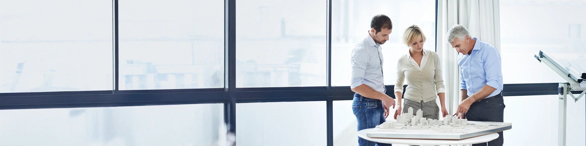 Tre professionisti, due uomini e una donna, sono riuniti intorno a un tavolo bianco all'interno di un ufficio luminoso con grandi vetrate sullo sfondo. L'uomo più anziano sulla destra sta indicando un plastico architettonico dettagliato di una città in miniatura appoggiato sul tavolo, mentre i colleghi lo ascoltano attentamente.