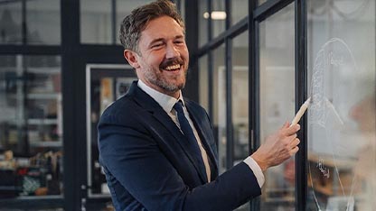 Uomo d'affari sorridente con la barba, che indossa un completo scuro e una cravatta, in piedi in un ufficio moderno con partizioni in vetro. L'uomo è girato verso destra, guardando verso una lavagna trasparente non visibile, e tiene un pennarello bianco nella mano destra come se stesse scrivendo o puntando qualcosa. La sua espressione è gioiosa e coinvolta. L'ambiente dell'ufficio sullo sfondo è leggermente sfocato ma mostra strutture in vetro e metallo.