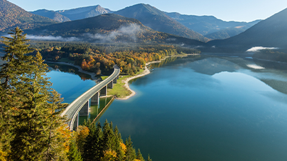 Una veduta panoramica dall'alto di un lago alpino circondato da montagne e boschi con colori autunnali. Un ponte stradale attraversa una parte del lago.