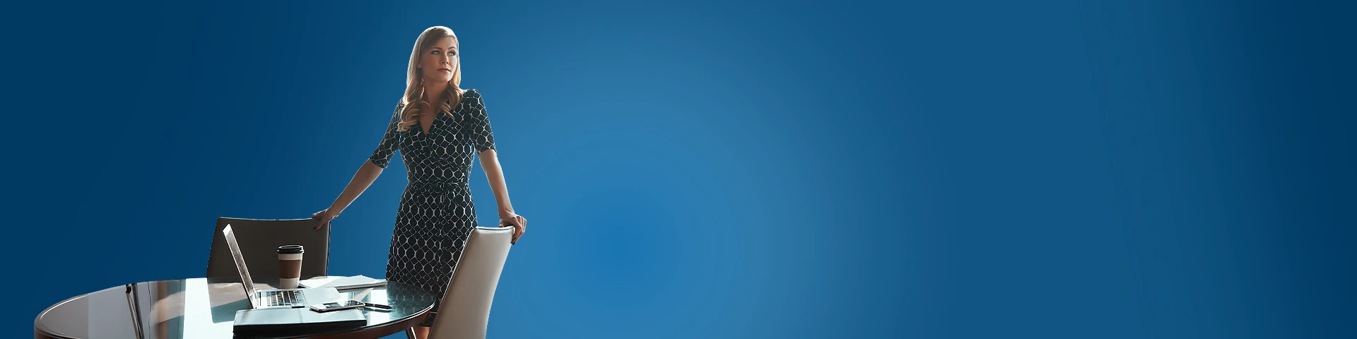 Una donna bionda in abito scuro a fantasia è in piedi dietro una scrivania con un laptop e una tazza di caffè. Guarda verso l'esterno dell'inquadratura con espressione attenta su uno sfondo blu sfumato.