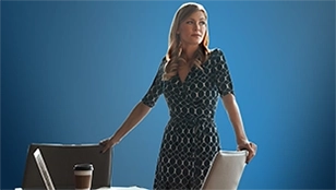 Una donna bionda in abito scuro a fantasia è in piedi dietro una scrivania con un laptop e una tazza di caffè. Guarda verso l'esterno dell'inquadratura con espressione attenta su uno sfondo di un grattacielo per cui alle spalle c’è la vetrata del palazzo