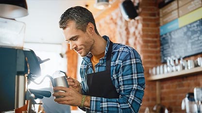 Interno di un bar moderno con pareti in mattoni a vista dove appare un barista. Lui è un uomo di mezza età, con capelli corti grigi e una camicia a quadri blu e bianca sotto un grembiule nero, è ritratto di profilo mentre utilizza una macchina per il caffè espresso professionale. È concentrato nel versare il latte montato da una lattiera in metallo in una tazza, creando una decorazione.