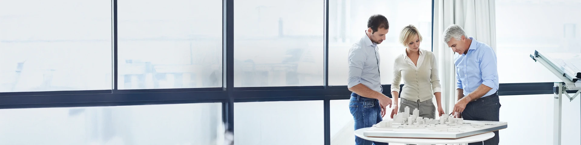 Tre professionisti, due uomini e una donna, sono riuniti intorno a un tavolo bianco all'interno di un ufficio luminoso con grandi vetrate sullo sfondo. L'uomo più anziano sulla destra sta indicando un plastico architettonico dettagliato di una città in miniatura appoggiato sul tavolo, mentre i colleghi lo ascoltano attentamente.