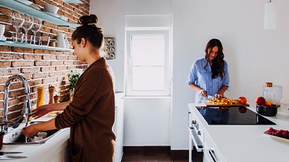 Giovane donna con capelli raccolti davanti al lavandino della cucina e giovane donna che affetta delle verdure su un tagliere
